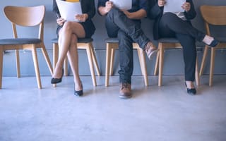 A row of chairs containing three individuals, visible from the neck down, in business garb and holding resumes.