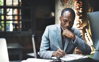 A man with salt-and-pepper hair in a gray business suit sits in contemplation at his desk in front of a dark office background.