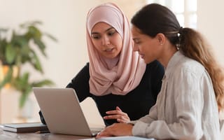 young arabian woman in hijab sitting with smiling colleague at table, looking at computer screen, explaining new company software