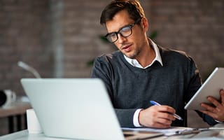 Man taking notes while holding a digital tablet and looking at a laptop