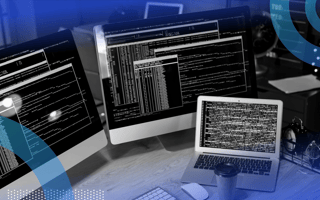 Two large screens, a laptop, and a to-go cup of coffee sit on a desk, displaying many commands in text boxes. /software-engineering-perspectives/programming-jobs-recession