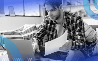 A young white man in a plaid shirt holds a document and types on a laptop. /product-management/must-document-no-code-low-code