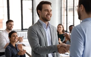 two employees in a meeting shaking heads with others clapping in the background