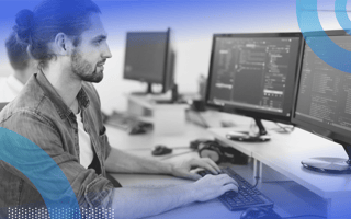 Ember.js image of a white man with a trimmed beard and his hair pulled back in a bun. He types code on a dual-monitor system. He wears a chambray shirt.