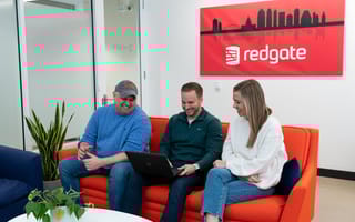 Three Redgate employees sitting on a red couch with a Redgate picture and logo behind them.