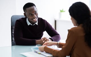 A young man and woman at a table. The man is smiling and talking to the woman, who is looking over some papers.
