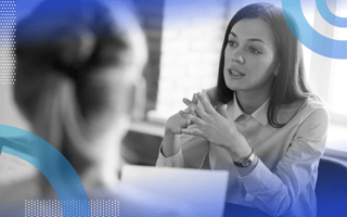 full cycle recruiting image of a woman holding her hands together in a kind of steeple pose while talking to people across a table. A woman's head is out of focus in the foreground and can be seen from behind.