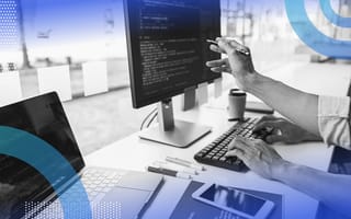 What Are JavaScript Design Patterns? JavaScript Design Patterns image of a person sitting at a computer monitor and a laptop. He holds a pen and points to something on the desktop screen. We can see code on both screens.