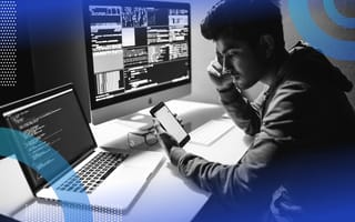 MVVM Architecture man sitting at his laptop and desktop computer. Both screens contain code. He is holding a cell phone