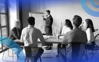 Stakeholder management image of a man in glasses presenting to a group of five people sitting around a table. The man looks at a large post-it and writes on it while all the people look at the man presenting.