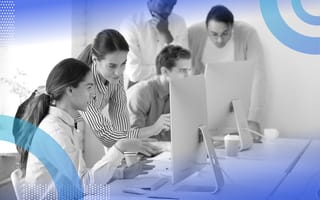 Supervised learning image of a group of professional people working together on a project. A man and a woman are in the foreground. He seeks her help with a problem. She stands next to him pointing at the screen.
