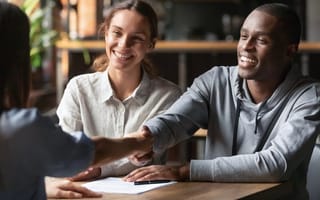 A Black man and woman smile while shaking hands with a salesperson across a table