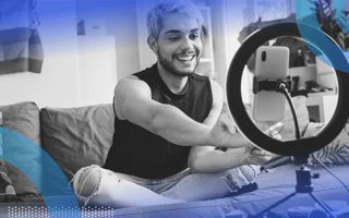 influencer marketing image of a person with bleach blonde hair and torn jeans sitting cross-legged on a couch. They reach forward to hit record on their smartphone, which sits inside a ring light.