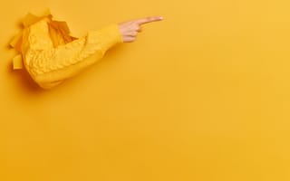 Faceless woman breaks arm through paper yellow wall and points.