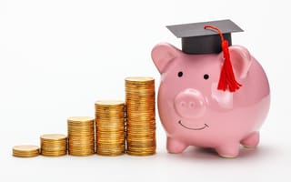 A graduation cap sits on top of piggy bank next to stacks of coins.