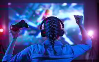 A gamer wears headphones and holds up a controller in front of a monitor.