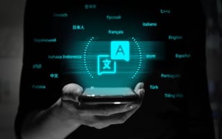 A hand holding a smart phone with glowing languages floating around it.