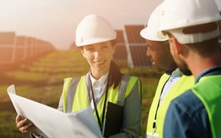 Three engineers wearing hard hats and work vests stand together looking over blueprints in a field surrounded by solar panels.