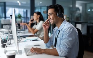 A tech support specialist types on a desktop computer and wears a headset while helping customers.