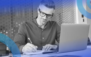 A man takes notes in a notebook while looking at a laptop