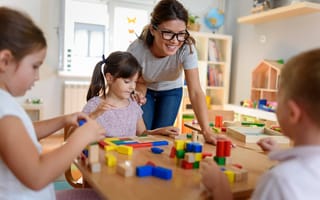A daycare teacher leans over a table of three children who are playing with colorful blocks.