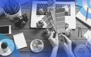 Hands hold color swatches in front of a laptop with a cup of coffee and wadded up balls of paper strewn about