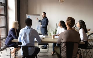 An instructor stands at the front of the room gesturing to a white board as he leads a training program for a group of professionals seated around a conference table.