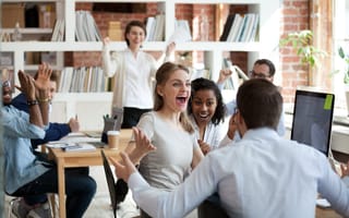 Several coworkers in a room together smile, clap and throw their hands in the air to represent employee engagement.