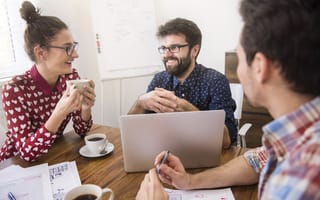 Three employees sit together at a table laughing, smiling and talking.