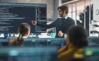 An instructor gestures to a large monitor containing lines of code as he leads a training course filled with people sitting at desktop computers.