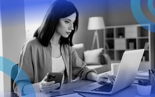A woman focuses on a laptop screen and uses a smartphone