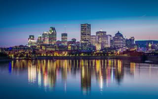 A view of the downtown Montreal skyline with several tall buildings lit up next to the water in early evening.