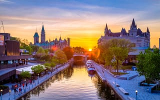 A view of the Ottawa skyline at sunset with large buildings in the background and people walking along a body of water.