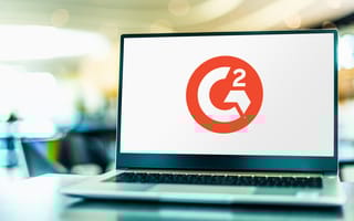 G2's red logo on a laptop. The laptop sits on a desk. A soft-focus office is visible in the background.