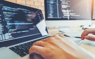 Close-up photo of software developer's hands on laptop keys with two computer screens in background showing code