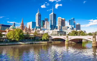 A view of the Melbourne skyline featuring a bridge over water in the foreground and several tall buildings in the back.