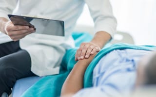 A doctor holds a clipboard while sitting next to a patient who is lying on their back.