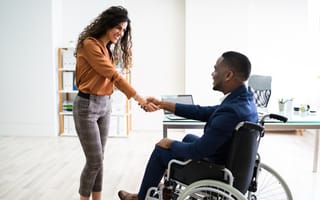 A businesswoman stands shaking hands with a businessman in a wheelchair.