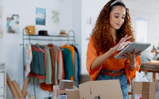 A woman with long curly hair stands in front of many open cardboard boxes holding a tablet. She wears a bright orange shirt and blue jeans with her long curly hair tied back in a stylish headband. A rack of clothing can be seen behind her as if she is in 