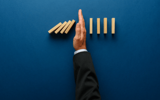 Hand in suit sleeve stops a row of falling wood blocks on a blue table