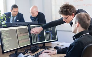 Two software professionals look over a pair of desktop computer monitors filled with lines of code.