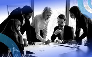A group of business people led by an older woman strategize at a table.