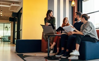 Young people (one male and three females) have a discussion while seated in a modern and bright office.