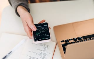 Person's hand holding a smart phone and looking at investment performance. They sit at a white desk with a gold macbook and a notebook.