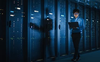 A person looking at an open laptop walks alongside network servers.