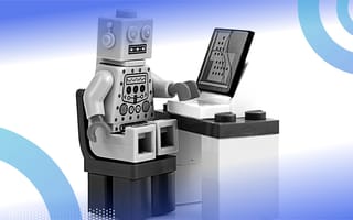 A toy robot seated at a desk and looking at a computer.