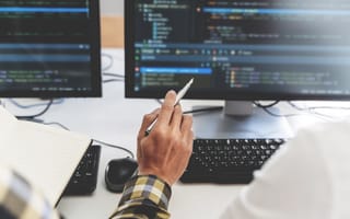 A close up of a hand holding a pen and gesturing to a computer monitor filled with lines of code.