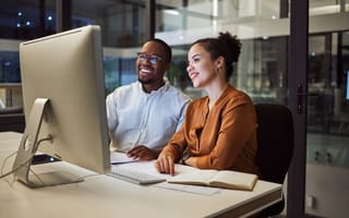 A pair of tech professionals sit together at a desk looking at a desktop computer monitor.