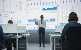 Zoom Out Back View Shot Of Female Data Scientist Using Laptop And Analyzing Information On Big Digital Screen in Monitoring Office. Diverse Employees Working On Desktop Computers For Big Data Company