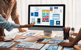 A development team brainstorming UI and UX designs for mobile apps. Only their hands are visible as they stand around a desktop computer screen.